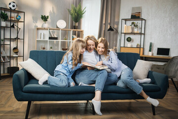 Canvas Print - Loving mother with two little kids reading book together, hugging, sitting on cozy couch at home, smiling mother with adorable two blond daughters engaged in educational activity