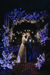 A stylish groom in a blue suit and a beautiful bride in a white dress hug near an arch decorated with flowers and lamps at a ceremony at night. Wedding photography, portrait of the newlyweds.
