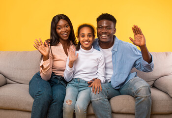 Wall Mural - Portrait of happy black man, woman and girl sitting on sofa, posing over yellow background and waving hands to camera