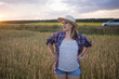 © olga_sova - A beautiful middle-aged farmer woman in a straw hat and a plaid shirt stands in a field of golden ripening wheat during the daytime in the sunlight