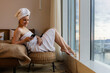 a young woman after shower in white bathrobe and towel on head sits on a bed in the morning at home and communicates by text messages on the phone. Rest in a hotel while traveling