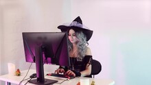 Young Beautiful Witch Wearing A Witch's Hat Sits At The Computer With Halloween Decorations, Typing On The Keyboard, Smiling And Waving Welcome, Come Closer, Pointing At The Computer Screen