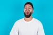 © Jihan - Stressful bearded caucasian man wearing white T-shirt over blue background screams in panic, closes eyes in terror, keeps hands on head, finds out terrified news, can't believe it.