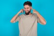 © Jihan - Happy bearded caucasian man wearing casual T-shirt over blue background ignores loud music and plugs ears with fingers asks to turn off sound