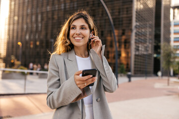 Wall Mural - Pretty young caucasian woman is using smartphone spending time outdoors. Brown-haired girl looks away, wears grey jacket. Technology concept