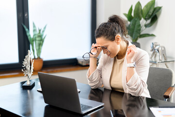 Depressed md-age woman feels tired sitting in front of a laptop in modern office, female employee massaging back of the neck, feels muscle strain. Overwork and overload concept