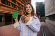 © Look! - Happy young caucasian girl pointing her finger at blank smartphone screen standing on street. Blonde with snow-white smile looks at camera, wears shirt. Technological device concept.