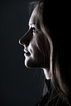 Close-up Woman Dark Profile Portrait In Low Light Studio Shot. Model Face Partly Illuminated By Light. Selective Focus And Image With Shallow Depth Of Field. Mysterious Atmosphere
