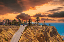 A Tall Brown Rocky Hillside At The Beach With A Long Street Down And Tall Lush Green Palm Trees And Plants With Powerful Clouds At Sunset At White Point Beach In San Pedro California USA