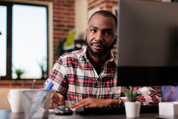 Wall Mural - African american man taking notes on files, writing report information on notebook at office desk. Working from home on startup documents, doing paperwork and browsing internet on computer.