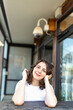 © elenarostunova - A smiling brunette girl in a white T-shirt, blue jeans sits at a wooden counter in a cafe, resting her chin in her hands. Vertical frame.