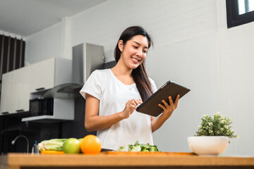 Wall Mural - Beautiful woman using tablet to calculate calories and nutrition vegetable salad in weight loss diet. Young female happily cooks a healthy breakfast in kitchen in the morning. Diet food concept.