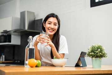 Wall Mural - Young female drinking water in glass for refreshing for diet and wellness on table. Beautiful woman happily drink fresh water before breakfast in kitchen in the morning. Healthy food concept.