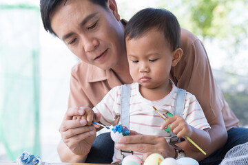 Father and baby little son painting eggs with brush in class workshop. Happy Asian dad embracing his little son and teaching painting eggs with water colour