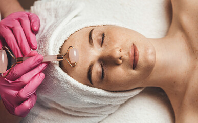Wall Mural - top view of a caucasian woman getting massage with jade face roller in spa salon. freckled face. cos