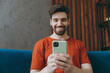 © ViDi Studio - Young fun man in red t-shirt hold in hand use mobile cell phone sit on blue sofa couch stay at home hotel flat rest relax spend free spare time in living room indoors grey wall People lounge concept