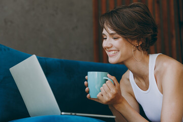 Side view young IT woman in white tank shirt hold cup use work on laptop pc computer lay down blue sofa stay at home flat rest relax spend free spare time in living room indoor People lounge concept