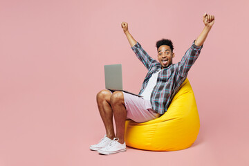 Wall Mural - Full body overjoyed happy young man of African American ethnicity wear blue shirt sit in bag chair hold use work on laptop pc computer do winner gesture isolated on plain pastel light pink background.