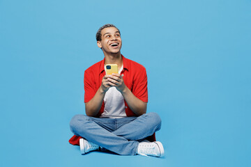 Wall Mural - Full body smiling young man of African American ethnicity 20s he wear red shirt sit in bag chair use mobile cell phone look aside on workspace area isolated on plain pastel light blue cyan background.