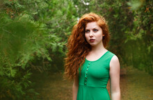Portrait Red Beautiful Young Woman, Against Green Summer Park. In Green Dress With Long Wavy Hair And Red Lipstick Posing