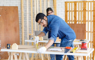 Wall Mural - Indian professional male engineer architect foreman labor worker wears safety goggles using measuring tape measure wood plank on working table while female colleague typing laptop computer behind