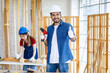 Asian Indian professional male engineer architect foreman labor worker wears safety hard helmet and gloves standing smiling holding thumb up and electric power drill machine in home construction site