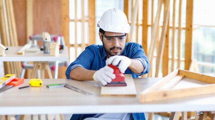 Wall Mural - Indian professional bearded male engineer architect foreman labor worker carpenter wears safety helmet and gloves using wood polishing machine grinding sanding wooden board surface on workbench