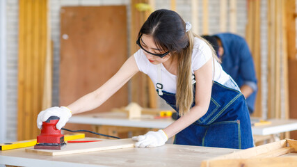 Wall Mural - Asian professional female engineer architect foreman labor worker wears safety goggles gloves using polishing machine grinding wood board on workbench while Indian male colleague use handsaw cutting