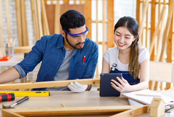 Wall Mural - Asian professional cheerful female engineer architect foreman labor worker sitting smiling holding tablet computer discussing with Indian bearded male carpenter in workshop housing construction site