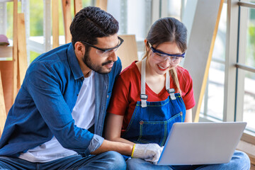 Asian professional female and Indian bearded male engineer architect foreman labor worker wear safety goggles and gloves sitting on floor smiling working with laptop computer discussing together