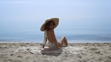 Wall Mural - Stylish woman turning back sitting sunny seashore. Curly girl sunbathing beach 