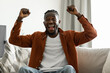 © Prostock-studio - Joyful black man cheering for sports team on television, gesturing YES and shouting, sitting on sofa at home