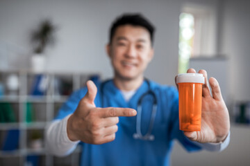 Wall Mural - Happy millennial korean male therapist pointing finger at jar of pills in clinic office interior. Treatment with doctor