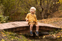 Little Blond Child Sitting On Bridge Over The Stream And Smiling In Warm Autumn Park With Golden Trees.