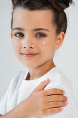 portrait of cheerful girl with lip gloss and sparkling glitter stars on face looking at camera isolated on grey.