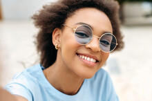 Modern Trendy Joyful African American Girl With Curly Hair, With Blue Glasses, Video Blogger, Takes A Selfie On The Cellphone, Looks At Frontal Camera, Records Online Stream, Smile Happily