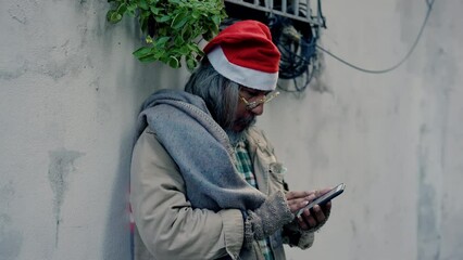 Wall Mural - 4K,  Homeless old Asian man wearing red christmas hat stands next to wall using his mobile phone to seek work and government assistance. Homeless people who do not have home to sleep on streets