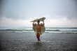 © Cavan Images - Man wearing angel wings carrying surfboard while walking in sea against cloudy sky