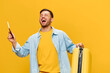 Joyful young tanned handsome man in blue shirt hold hands with passport plane tickets and suitcase up posing isolated on yellow studio background. Copy space Banner Mockup. Trip journeys concept