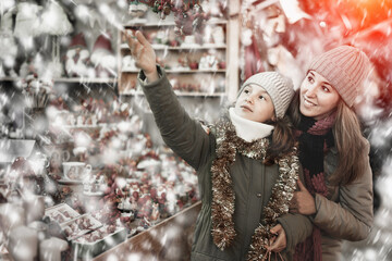 Positive female and her daughter are choosing decorations for Christmas tree in the market outdoor.