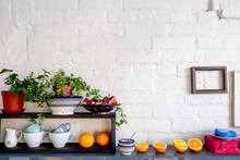 Potted Plants And Fruits With Kitchen Utensils On Table Against Brick Wall