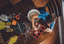 High Angle View Of Boy Putting Muffin Batter Into Baking Sheet's Containers On Table At Home