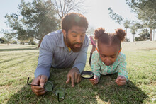 Father And Daughter Playing With Magnifying Glass While Lying On Grassy Field At Park During Sunny Day