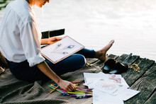 Low Section Of Woman Drawing While Sitting On Pier Over Lake During Sunset