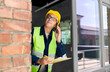 © Syda Productions - construction business and building concept - happy smiling male builder in helmet and safety west with clipboard calling on smartphone at window