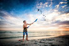 Full Length Of Shirtless Boy Throwing Sand In Air With Shovel At Beach Against Cloudy Sky During Sunset