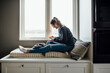 © Cavan Images - Side view of woman using mobile phone while sitting on alcove window seat at home