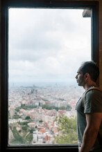 Side View Of Man Looking At Cityscape Against Cloudy Sky Through Window While Standing In Hotel Room