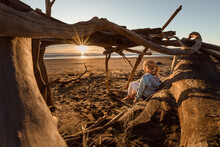 Blonde Girl Sitting In A Driftwood Hut Watching The Sunset At A Beach