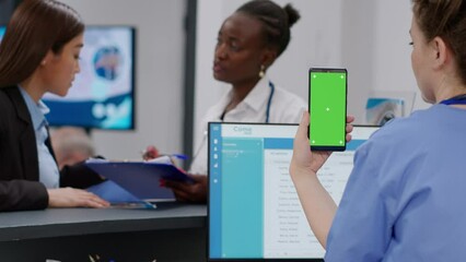 Medical assistant holding smartphone with greenscreen display at hospital reception counter. Nurse using chroma key template with isolated mockup background and blank copyspace.